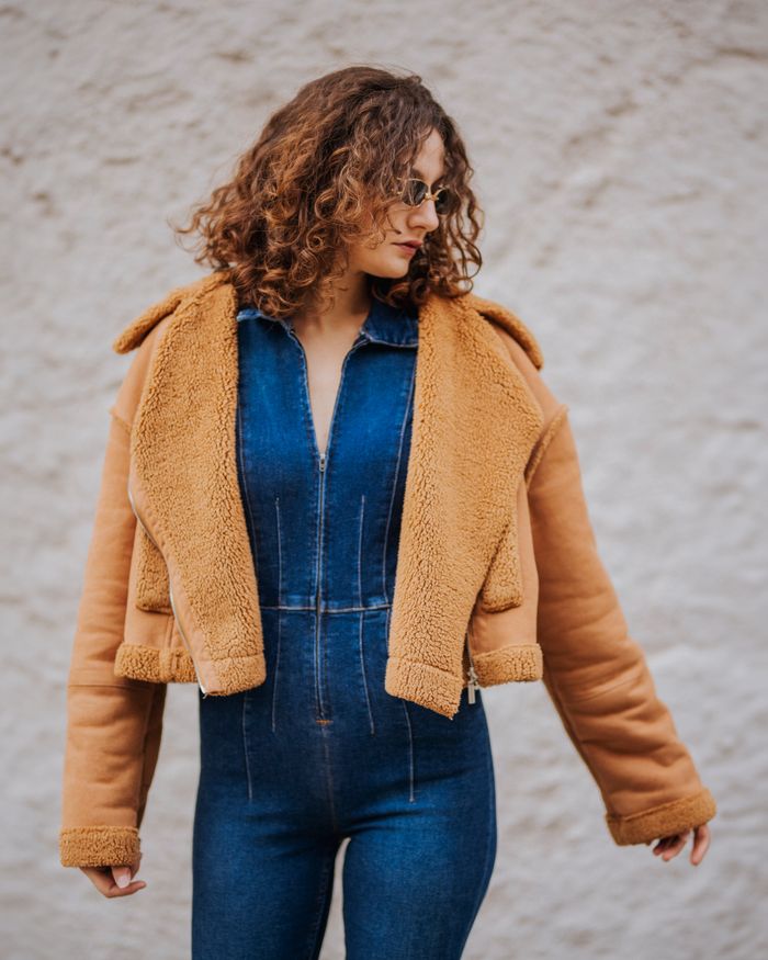 services-04 Fashion portrait of a woman in sunglasses, wearing a denim jumpsuit and brown jacket against a textured wall.